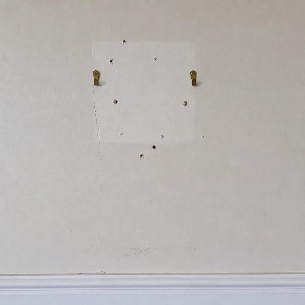 Close-up of brass picture hooks, nail holes and a cleaner rectangle of paint on an interior wall