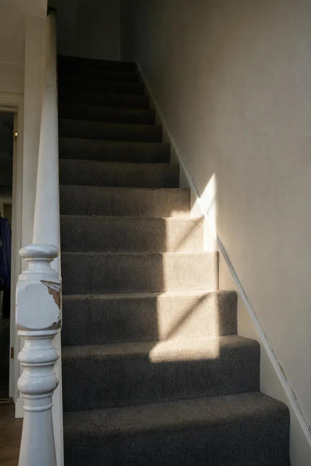 Afternoon light falls down a narrow domestic staircase with a worn grey carpet runner